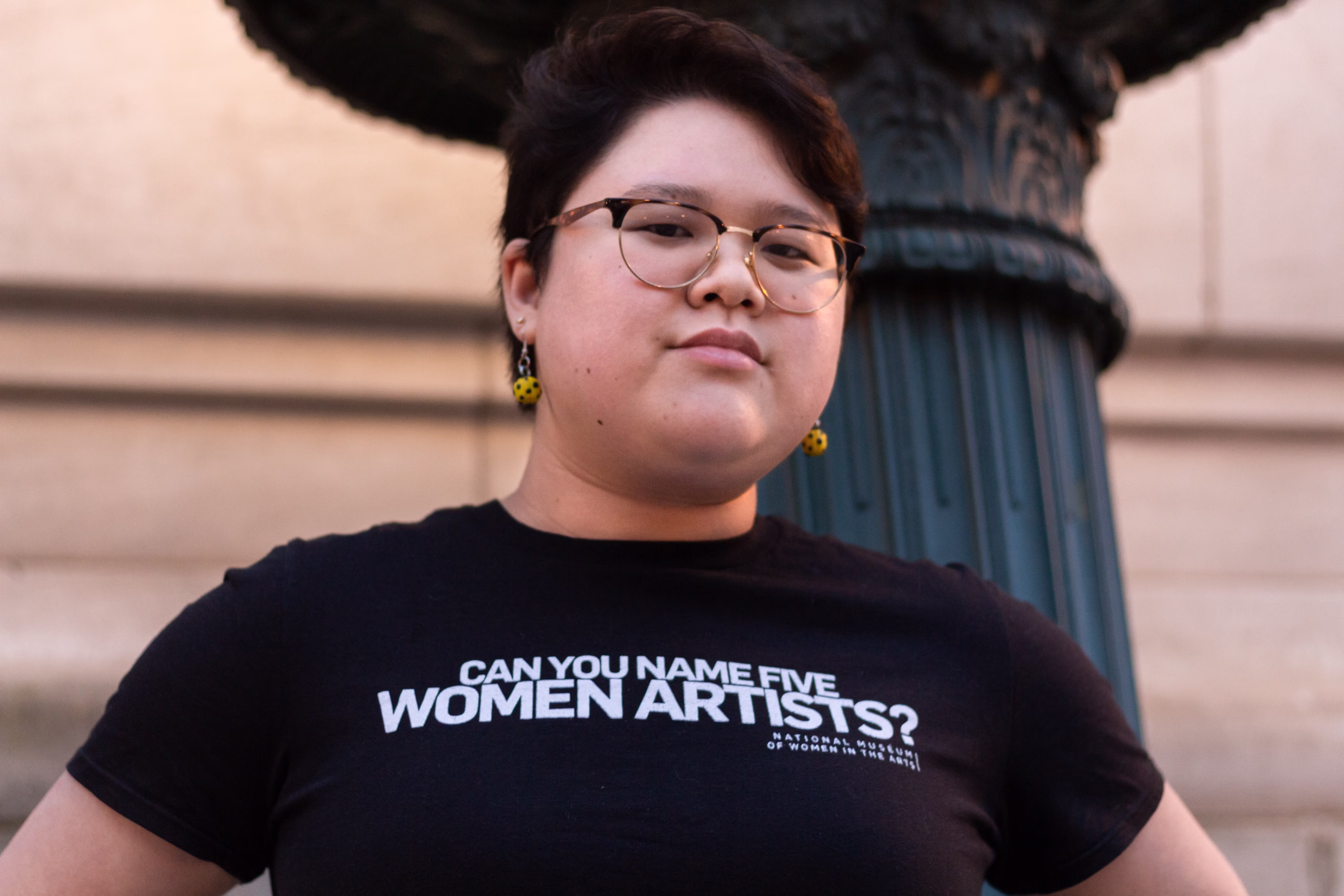 A young woman of Asian descent poses in a black T-shirt that has the question "Can you name five women artists?" printed in bold white text in its center.