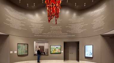 A museum rotunda features a red chandelier hanging from the center of the ceiling. The chandelier is made of red and orange glass and festooned with ornamented red-and-gold fabric garlands. LED lighting around the glass bulbs illuminates the chandelier.