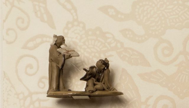 A woman wearing an elaborate hairstyle and earrings is seen in profile against a cream background and outlines of butterflies. A decorative glass bottle and two miniature ceramic sculptures of male figures are attached to the surface of the print.