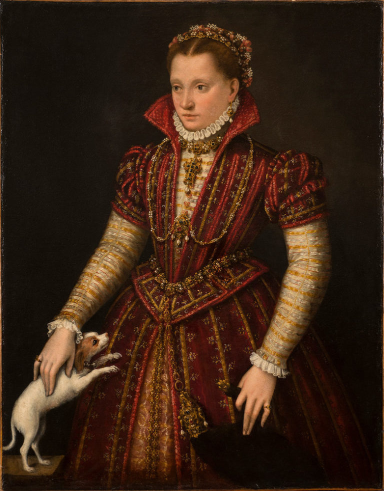A woman stands in a richly brocaded red dress, her right hand reaching down to pet a small, white dog. Adorned in jewelry, she wears flowers in her hair, parted in the center. The pelt of a small mammal, its head encased in a jeweled holder, hangs from her heavily decorated belt.