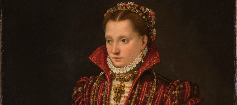 A woman stands in a richly brocaded red dress, her right hand reaching down to pet a small, white dog. Adorned in jewelry, she wears flowers in her hair, parted in the center. The pelt of a small mammal, its head encased in a jeweled holder, hangs from her heavily decorated belt.