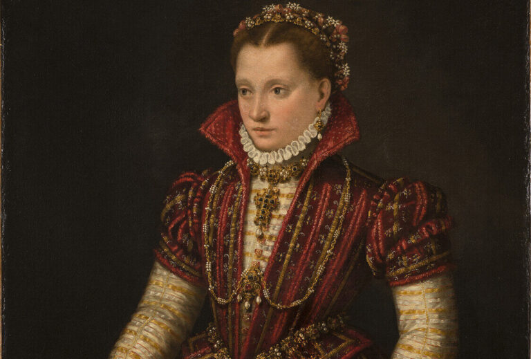 A woman with light skin tone stands in a richly brocaded red dress, her right hand reaching down to pet a small, white dog. Adorned in jewelry, she wears flowers in her hair, parted in the center. The pelt of a small mammal, its head encased in a jeweled holder, hangs from her heavily decorated belt.