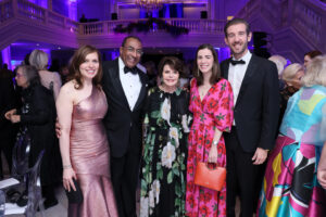 Three women and two men, all in evening wear, pose for a picture together inside of a large marble hall.