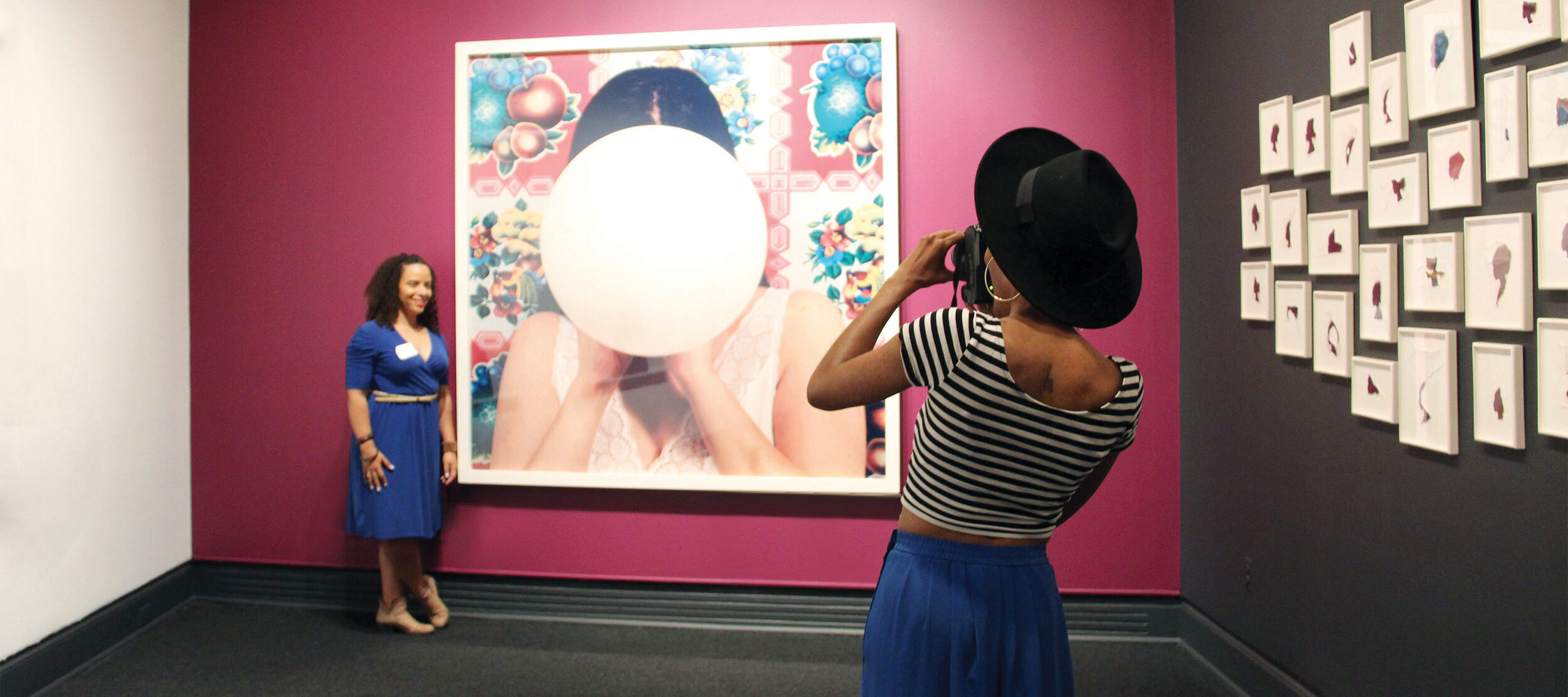 A woman in a blue dress poses next to a large, colorful artwork on a magenta wall, while another person in a striped shirt, blue skirt, and black hat takes her photo.