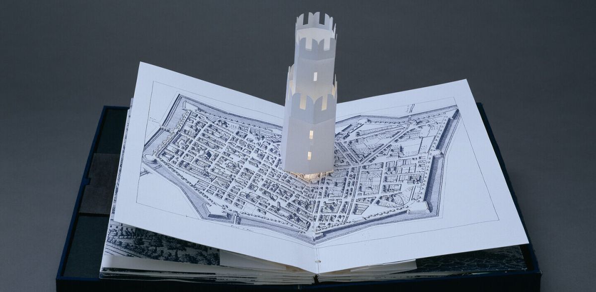 Bound in a dark blue fabric-covered binding, the book opens to display five pop-up towers illuminated from within by a tiny bulb. The featured page includes a white country church with a steeple, set into pages featuring musical scores presented in manuscript style.