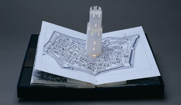 A pop-up book displays an intricate paper model of a tower rising from a map illustration of an old city, all set against a dark gray background.