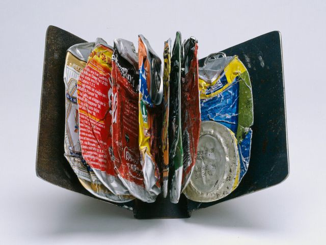 A collection of crushed and flattened aluminum beverage cans displayed in a manner resembling the pages of an open book. The cans are various colors, including red, yellow, and silver, and are arranged vertically between two black, book-like ends.