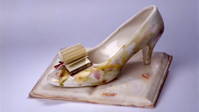 A woman's glass slipper rests on the open pages of a porcelain book. In place of a bow on the toe of the slipper is a paper book with pages fanned out.