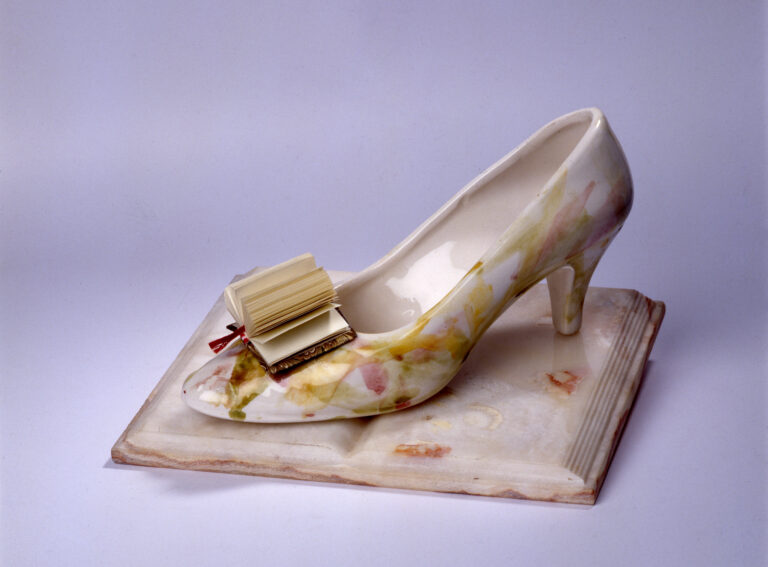 A woman's glass slipper rests on the open pages of a porcelain book. In place of a bow on the toe of the slipper is a paper book with pages fanned out.