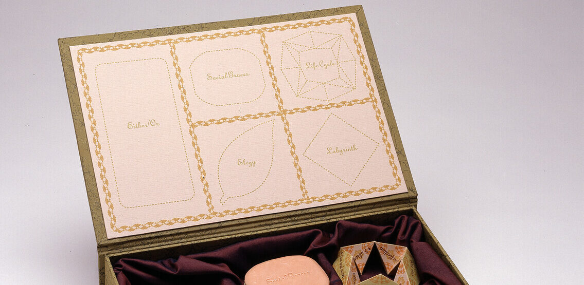 A green-gold box with its attached lid flipped open holds five objects shaped like candies resting on burgundy fabric. The inside of the lid displays outlines of each object, with the words "life cycle," "labyrinth," "elegy," "either/or," and "social graces" inside the outlines.