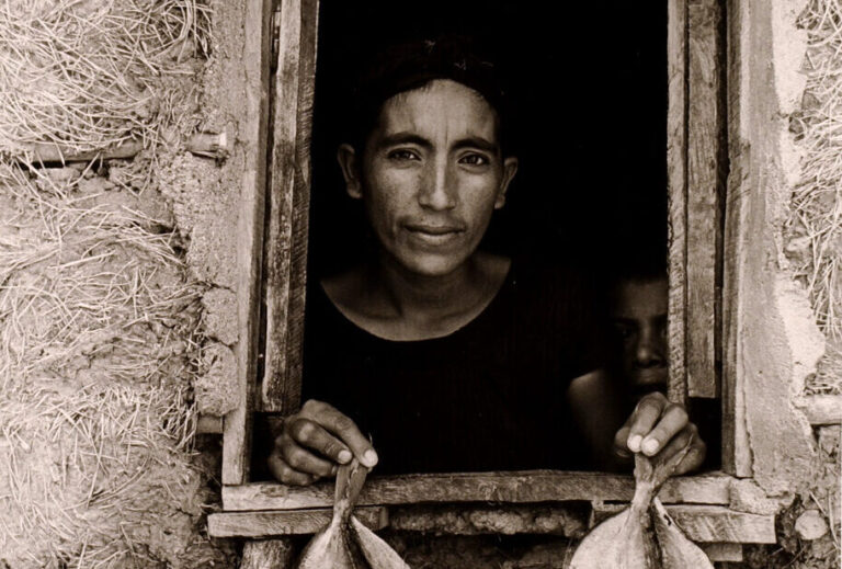 A black-and-white photograph features an open, rough-hewn window framing the head and shoulders of a woman with dark hair and medium-dark skin. Peering out from a murky interior, she displays 2 fish per hand on the window ledge. A mud-grass mixture textures surrounding walls.