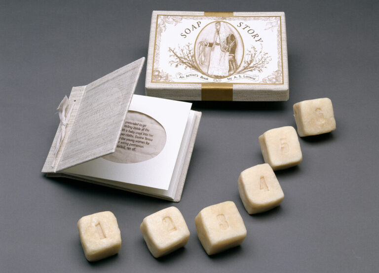 A set of five ivory-colored soap bars numbered 1 to 5 is arranged in a line in front of an open booklet and a box labeled "Soap Story," all displayed on a gray surface.