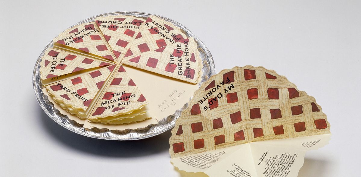 A pie tin filled with paper that resembles a whole pie with a lattice top and red filling. In black ink, the words “Eight Slices of Pie” is written across the crust, and “By Emily Martin” written in smaller text underneath. The pie tin is covered with a clear plastic top.