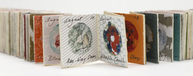 An accordion-style artist’s book is open, displaying pages with colorful abstract circular designs and handwritten notes, including dates in August. The book stretches out in both directions against a white background.