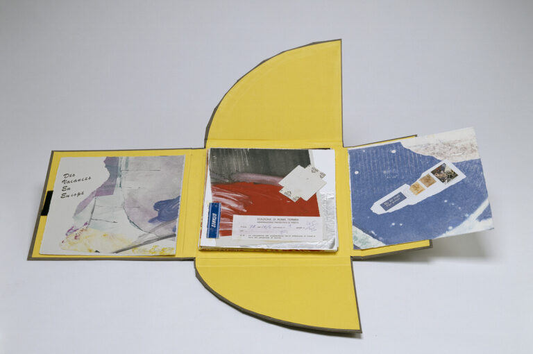 A yellow, book-like folder with rounded flaps is open, revealing abstract artwork, handwritten notes, and small printed items arranged neatly inside. The background is plain white.