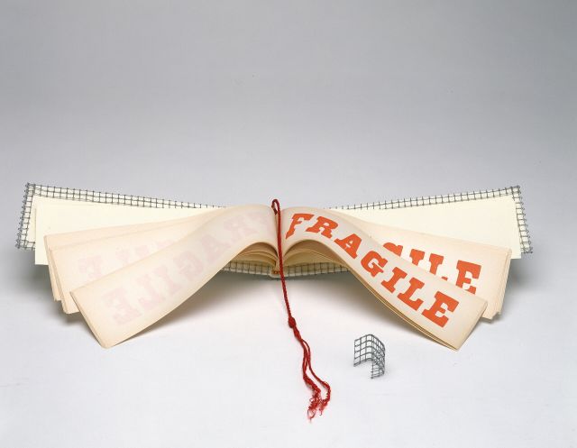 A wide and narrow book opened to display pages with the word FRAGILE in red block letters. A red tassel hangs down the center of the book and the cover is wrapped with wire mesh.