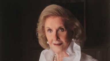 Professional portrait of a light-skinned, older woman with a blonde bob. She wears an elegant white ruffled blouse and pearl jewelry.