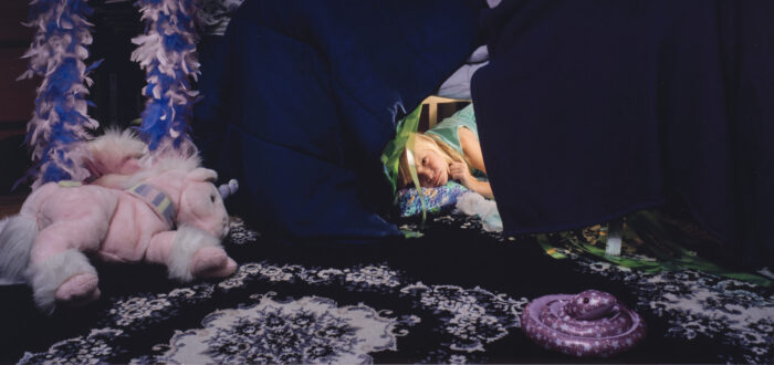 A color photograph of a young light skinned girl with light blonde hair. The girl peeks out from a fort made from a dark blue and purple comforter. A light pink and purple stuffed unicorn sits on a purple and white floral rug in front of the fort. A purple, blue, white, and black paper butterfly-shaped kite hangs on the wall above the fort.