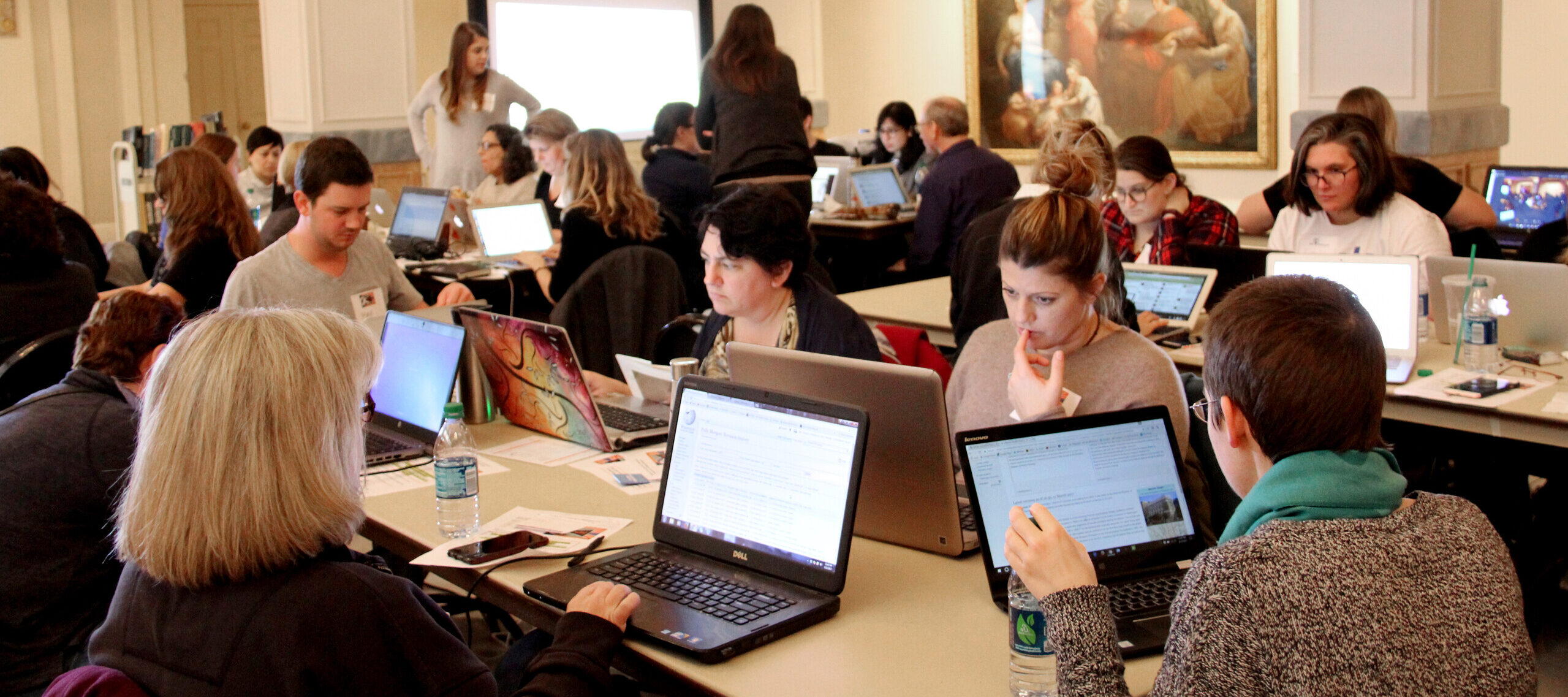 Multiple people are seated around rectangular tables working on laptops.