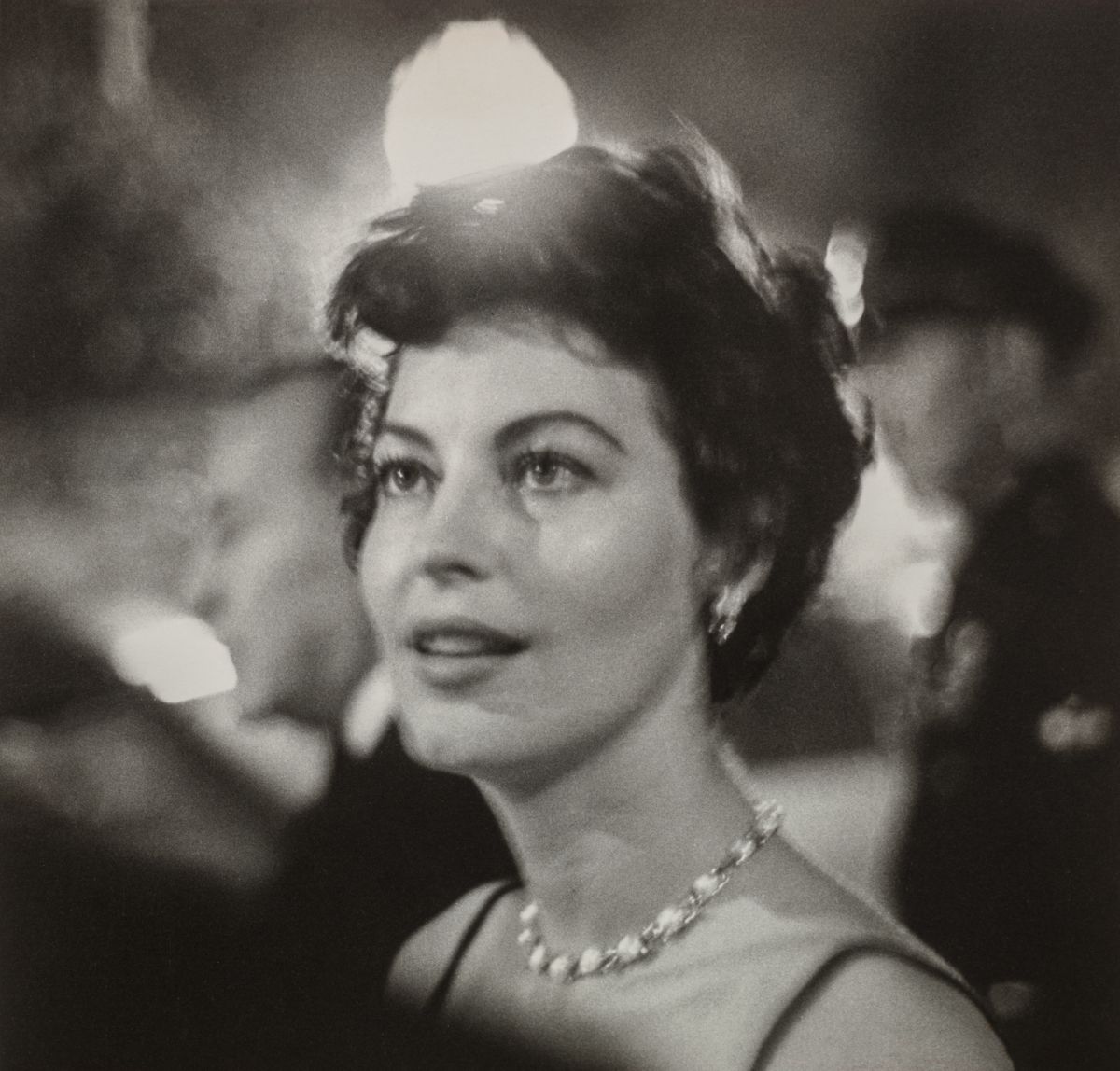 Ava Gardner, a woman with light skin tone and wavy, short brown hair, is shown from the shoulders up. She wears a jeweled necklace and is surrounded by lights and people at a party. She looks to the side of the camera at something out of frame with a fixed gaze and slight smile.