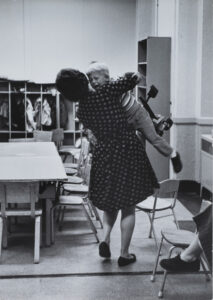 A black and white photograph taken from behind as a woman holds a crying child in a classroom setting.