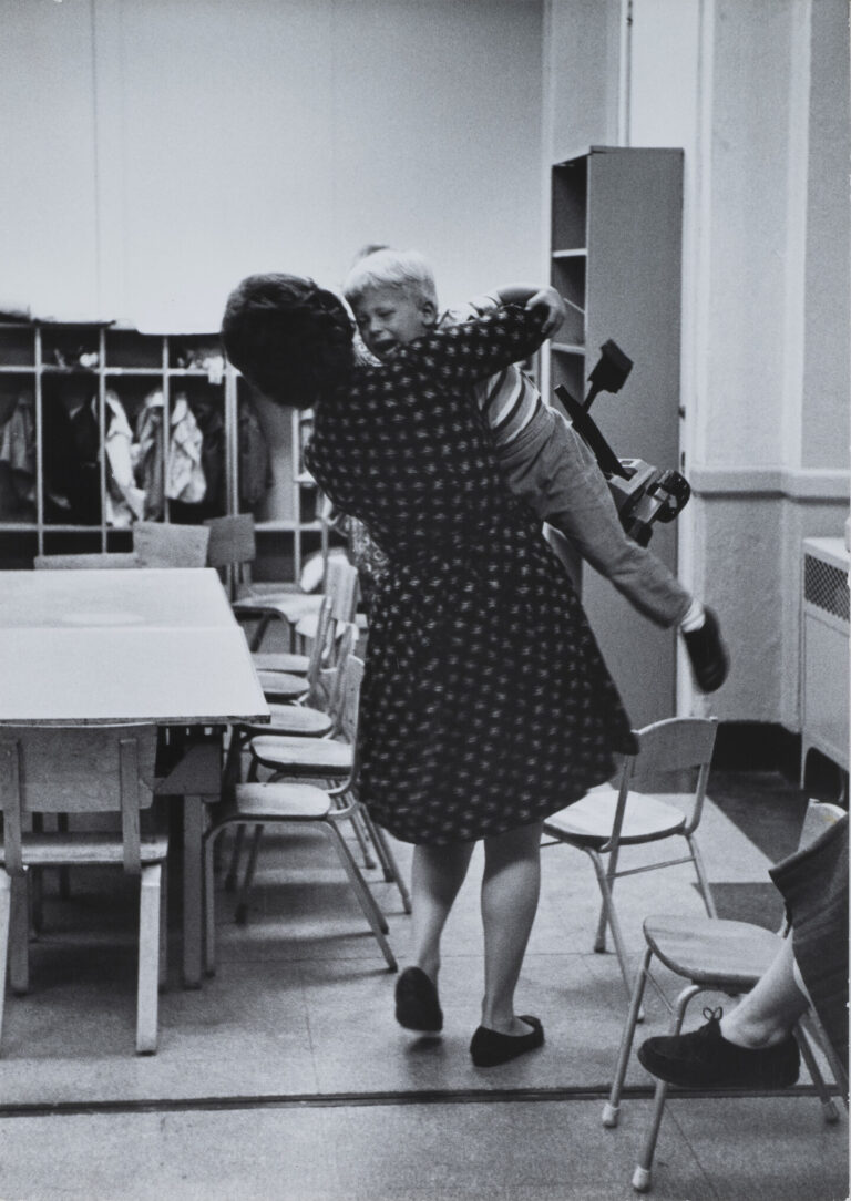 A black and white photograph taken from behind as a woman holds a crying child in a classroom setting.