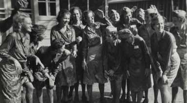 A black and white photograph of 14 women in matching uniforms, all of which have skirts covered in mud. Some women wear matching caps. Some women are touching the bottoms of their dirty skirts.
