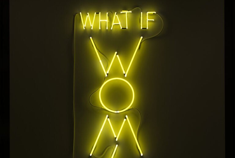 The phrase 'What if Women Ruled the World' is sculpted with neon tubes of bright yellow, in all capital letters. The letters of “WOMEN” are larger than the rest and are configured vertically.