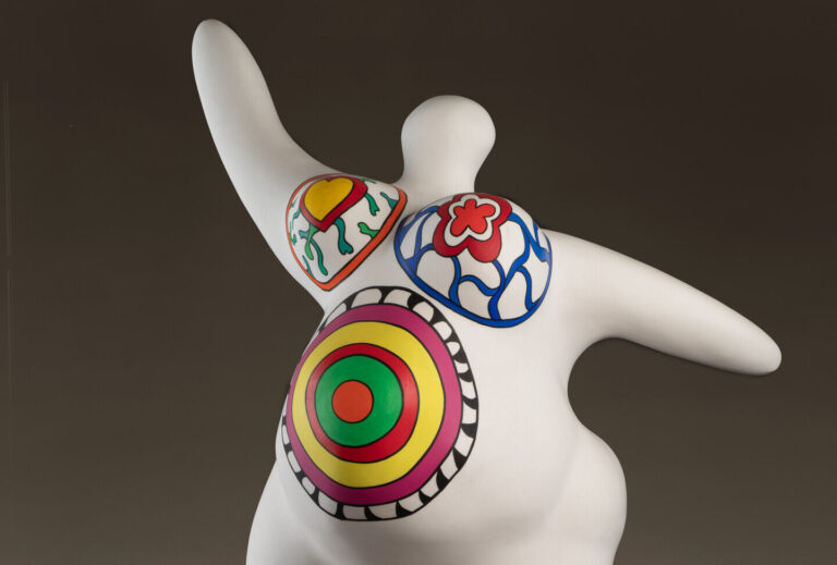 A marble sculpture depicts an abstracted, voluptuous figure with a pregnant belly. The figure is covered in bright patterns and posed stepping forward, with raised, outstretched arms.