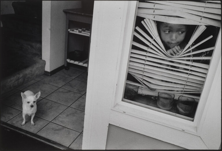 A black-and-white photograph of a young, medium-dark skinned girl peeking out between white blinds in a window that she bends and holds open with one hand. To the left of the window is an open doorframe to a tiled room where a little white dog stands.