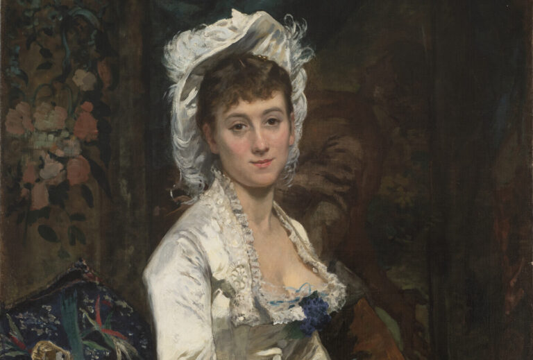 A light-skinned woman with brown hair wears an ivory gown and matching hat with a blue flower at the dress neckline. She is sitting with her hands in her lap, looking at the viewer with a pleasant smile.