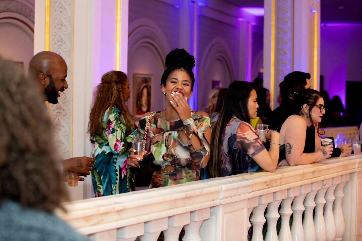 A group of people stand along an ornate white balcony at a lively indoor event. One woman in the center smiles and laughs, holding a drink, while others around her chat and enjoy the atmosphere.