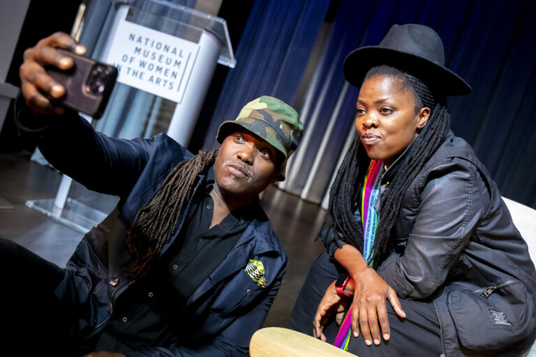 Two people with long braided hair take a selfie together indoors. One wears a camouflage hat and dark clothes, the other wears a black hat and colorful scarf. A podium labeled "National Museum of Women in the Arts" is in the background.