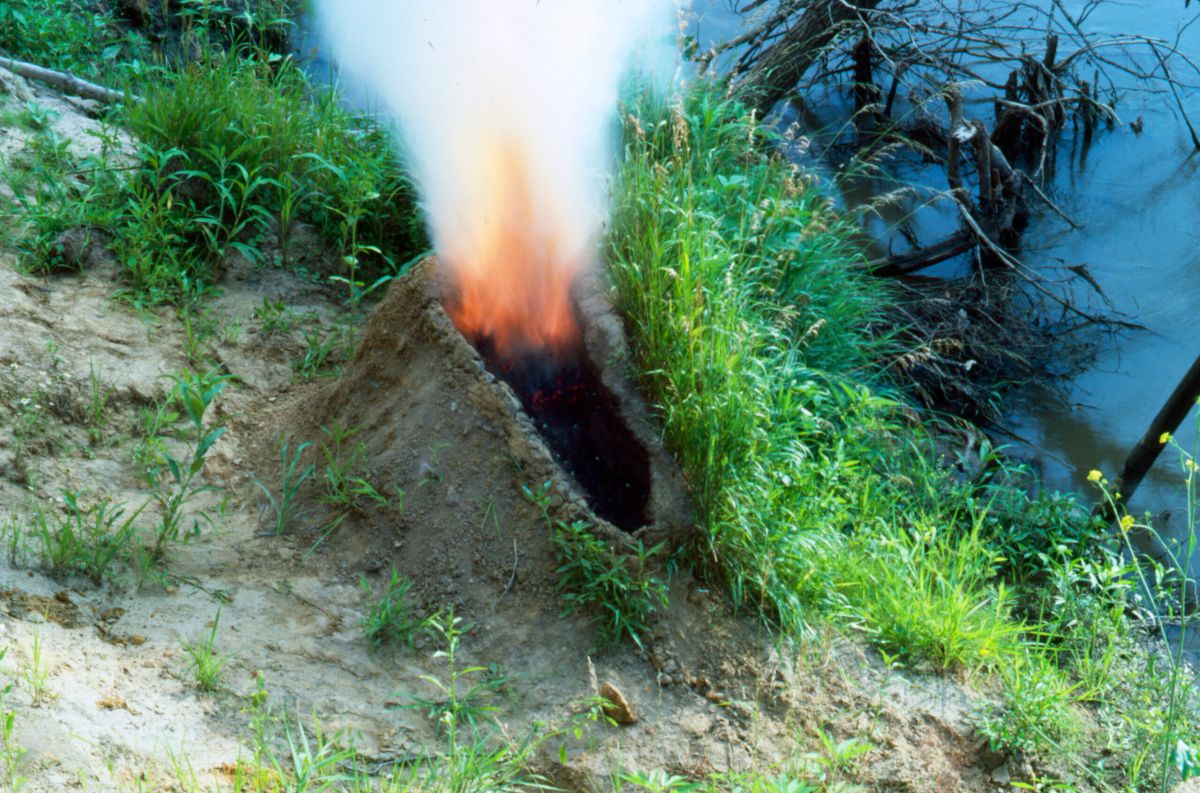 A mound of dirt in a grassy area by water. In the center of the mound is a human-shaped crater full of gunpowder that has been set aflame, exploding up in fire and smoke, making the dirt mound resemble an erupting volcano.
