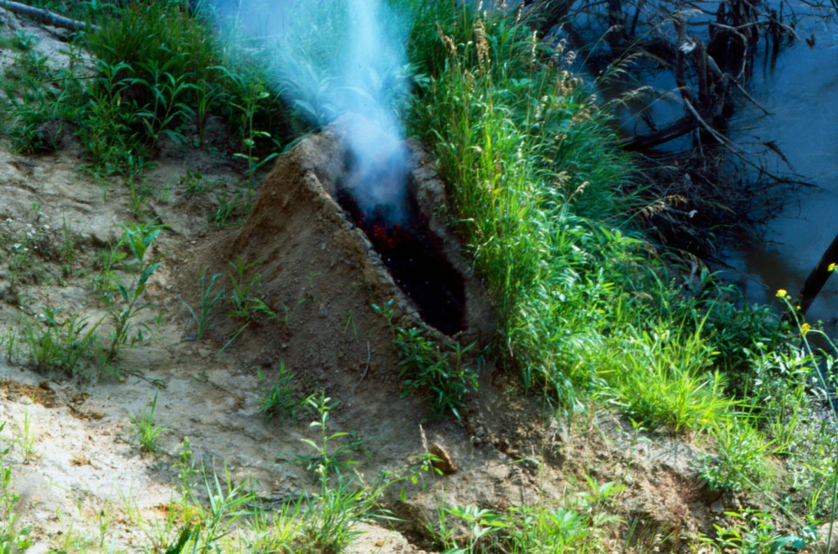 A mound of dirt in a grassy area by water. In the center of the mound is a human-shaped recess full of gunpowder that has been set aflame, exploded, and now the remaining ashes billow smoke into the air.