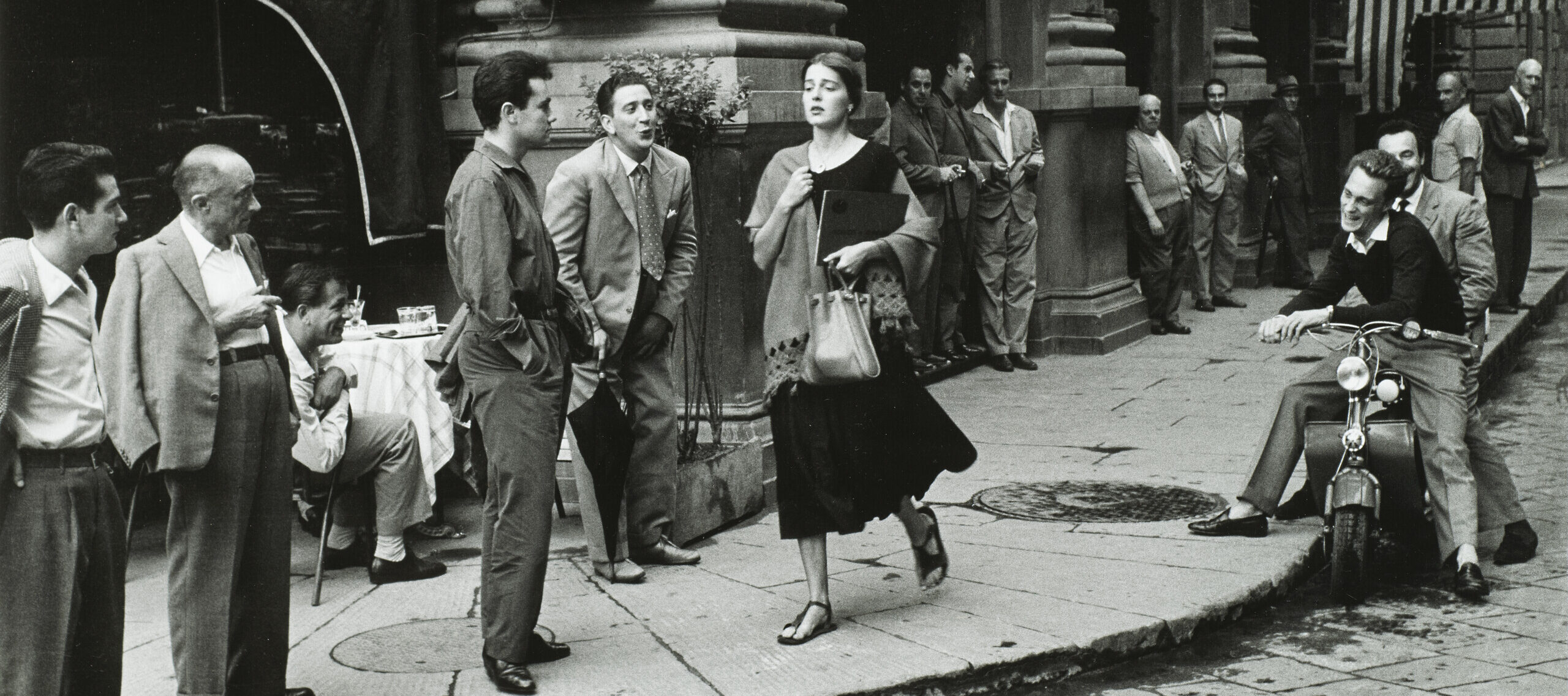 A black-and-white photograph of a young woman with light skin tone and brown hair wearing a black dress and a scarf and clutching a book and a bag walks down a city sidewalk. Several men watch her walk by, some standing against the wall of a nearby building, some sitting on chairs or motorcycles. A few men seem to be catcalling her.