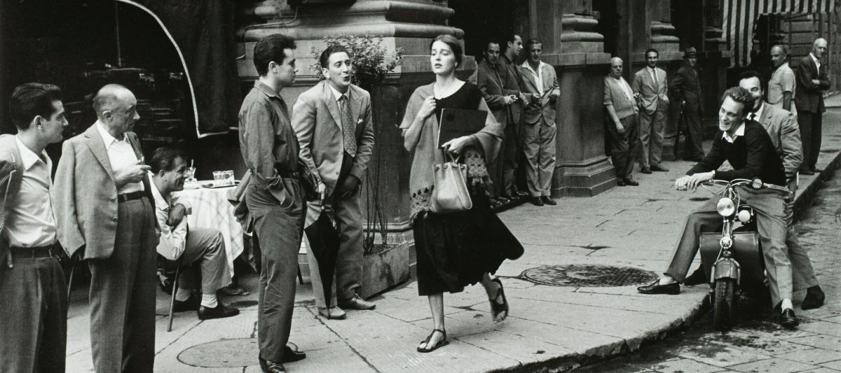 A black-and-white photograph of a young woman with light skin tone and brown hair wearing a black dress and a scarf and clutching a book and a bag walks down a city sidewalk. Several men watch her walk by, some standing against the wall of a nearby building, some sitting on chairs or motorcycles. A few men seem to be catcalling her.