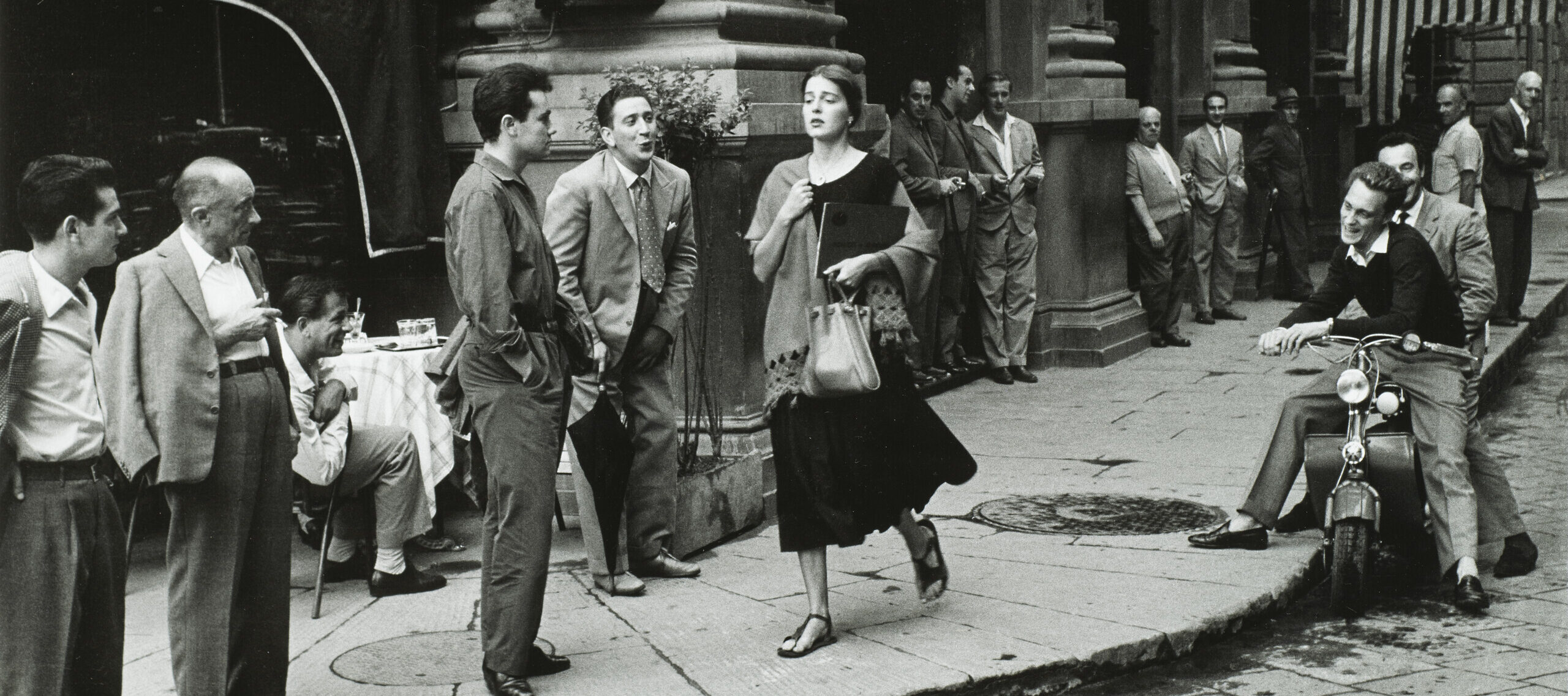 A black-and-white photograph of a young woman with light skin tone and brown hair wearing a black dress and a scarf and clutching a book and a bag walks down a city sidewalk. Several men watch her walk by, some standing against the wall of a nearby building, some sitting on chairs or motorcycles. A few men seem to be catcalling her.