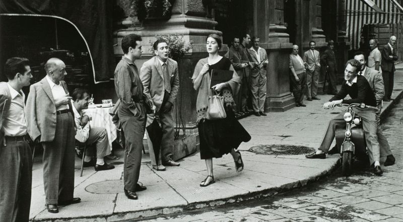 A black-and-white photograph of a young woman with light skin tone and brown hair wearing a black dress and a scarf and clutching a book and a bag walks down a city sidewalk. Several men watch her walk by, some standing against the wall of a nearby building, some sitting on chairs or motorcycles. A few men seem to be catcalling her.