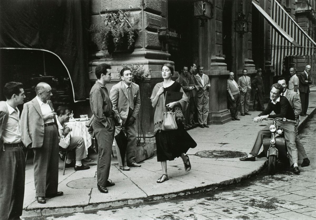 A black-and-white photograph of a young woman with light skin tone and brown hair wearing a black dress and a scarf and clutching a book and a bag walks down a city sidewalk. Several men watch her walk by, some standing against the wall of a nearby building, some sitting on chairs or motorcycles. A few men seem to be catcalling her.