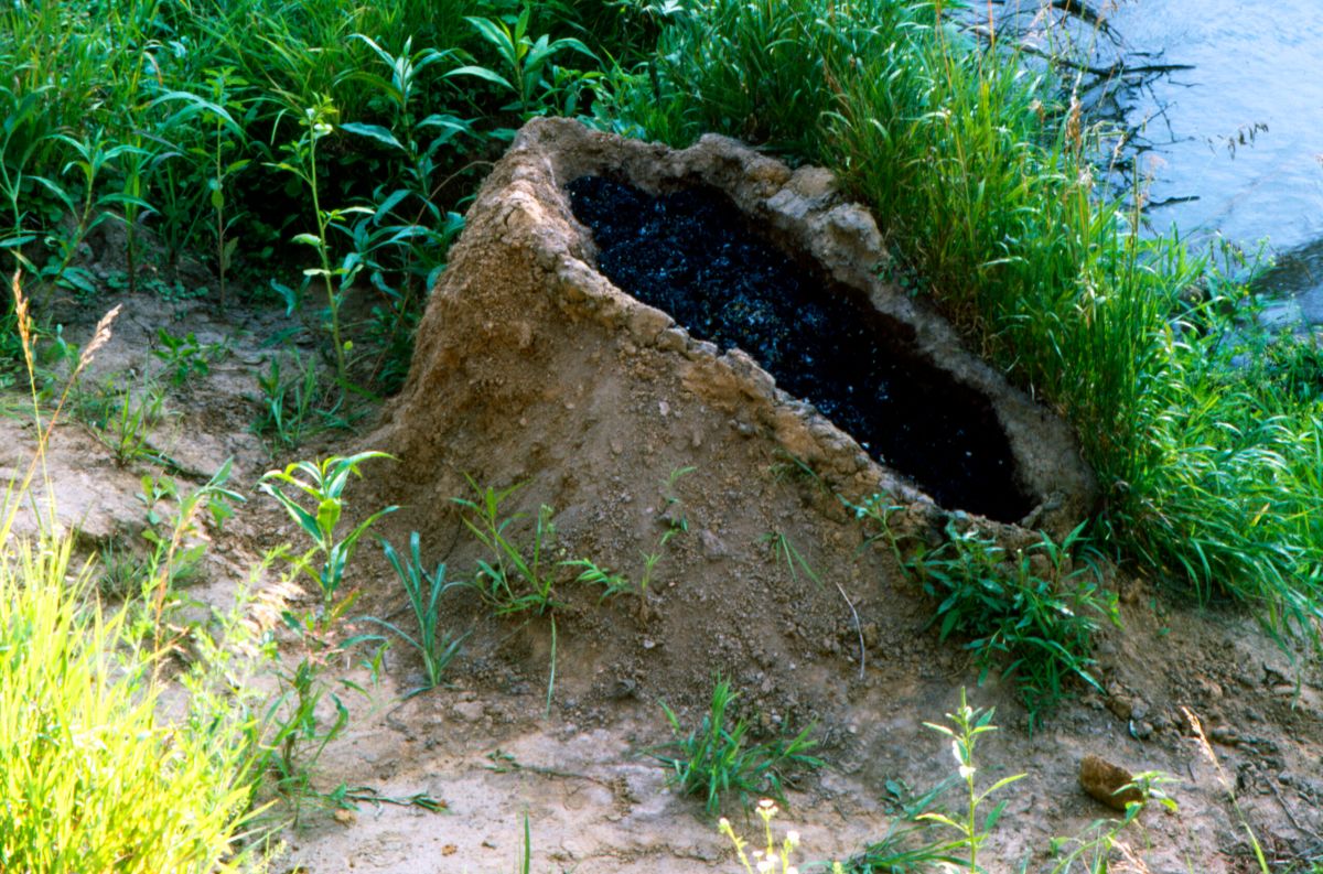 A mound of dirt in a grassy area by water. In the center of the mound is a human-shaped recess full of gunpowder that has been set aflame, exploded, and now is filled with black lifeless ashes.