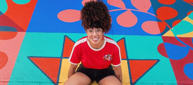 A young woman with coily, curly hair worn in a high ponytail sits in the middle of a bright mural painted on the ground. She smiles happily.