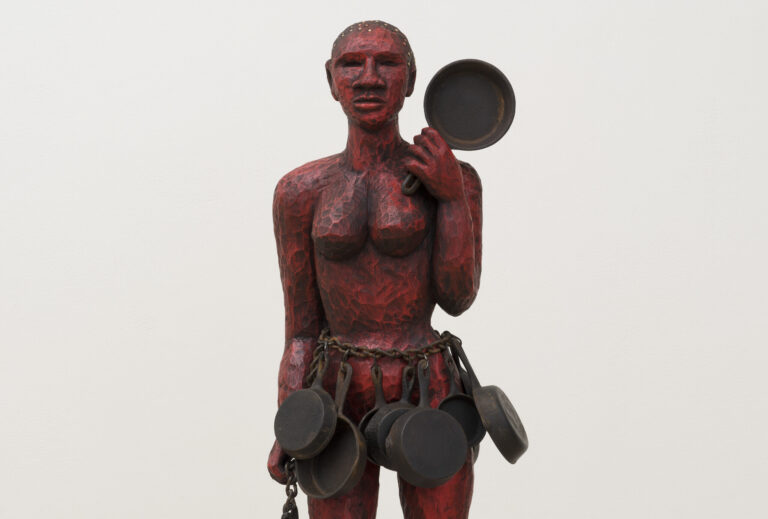 A red painted wooden sculpture of a woman holding a cast iron skillet in her left hand. She has a heavy chain around her waist with multiple skillets hanging from it like a skirt. She stands on a small wooden platform.