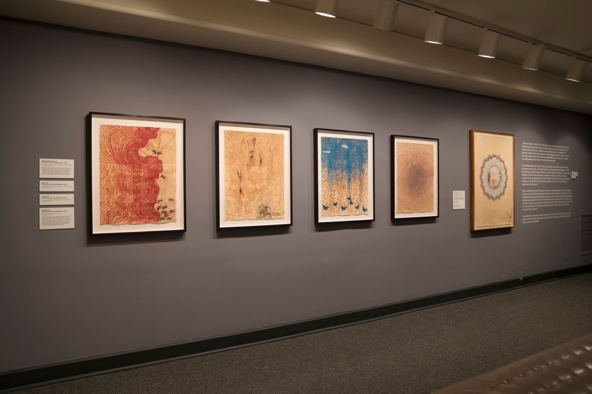 View of a gallery with dark walls. Bright works on paper are hanging on the walls.
