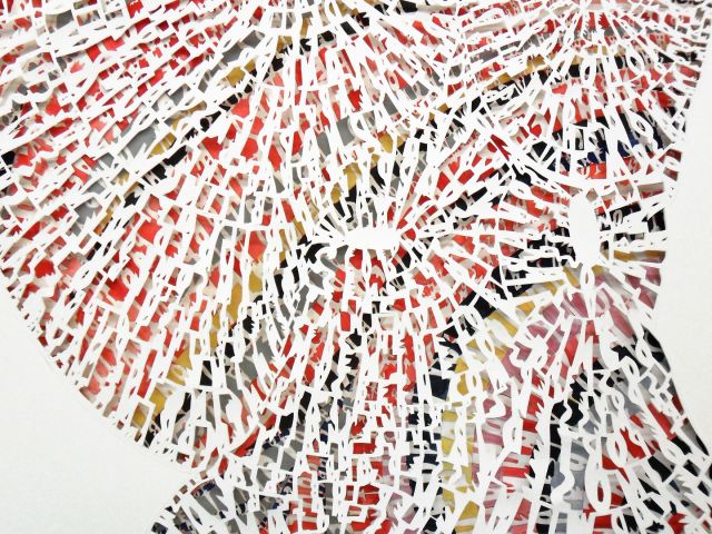 Close up of an intricately cut paper work. Under layers are vibrantly colored, with a layer of white paper with intricately cut alphabet pieces revealing the color below.