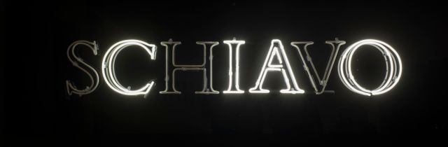White, capital, neon letters spell the Italian word 'schiavo' in serif text against a black background. The 's,' 'h,' and 'v' are dark, leaving the letters to spell 'ciao.'
