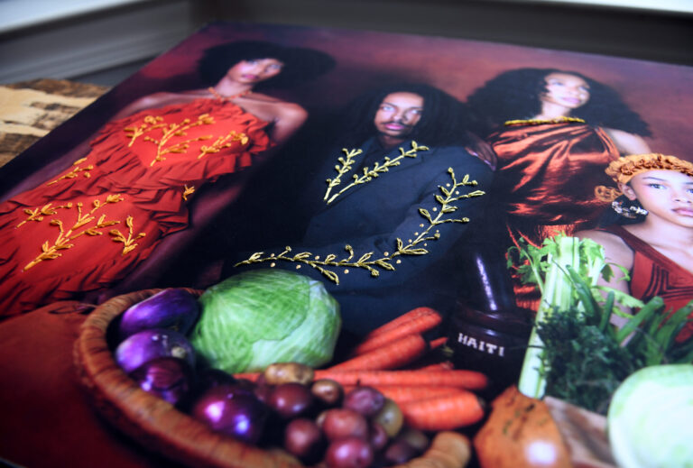 An artwork in process features figures standing behind a bowl of vegetables. Shown in a close, angled view, the artwork is a photograph with gold-colored thread stitched onto the figures’ clothing.