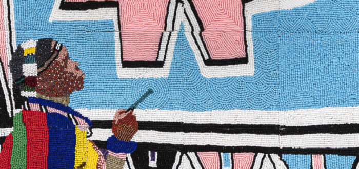 Tiny, colorful beads arranged in patterns to depict Esther Mahlangu painting a large artwork of geometric shapes in light pink and blue, black, white, yellow, and green. She is a dark-skinned adult woman wearing colorfully patterned robes.