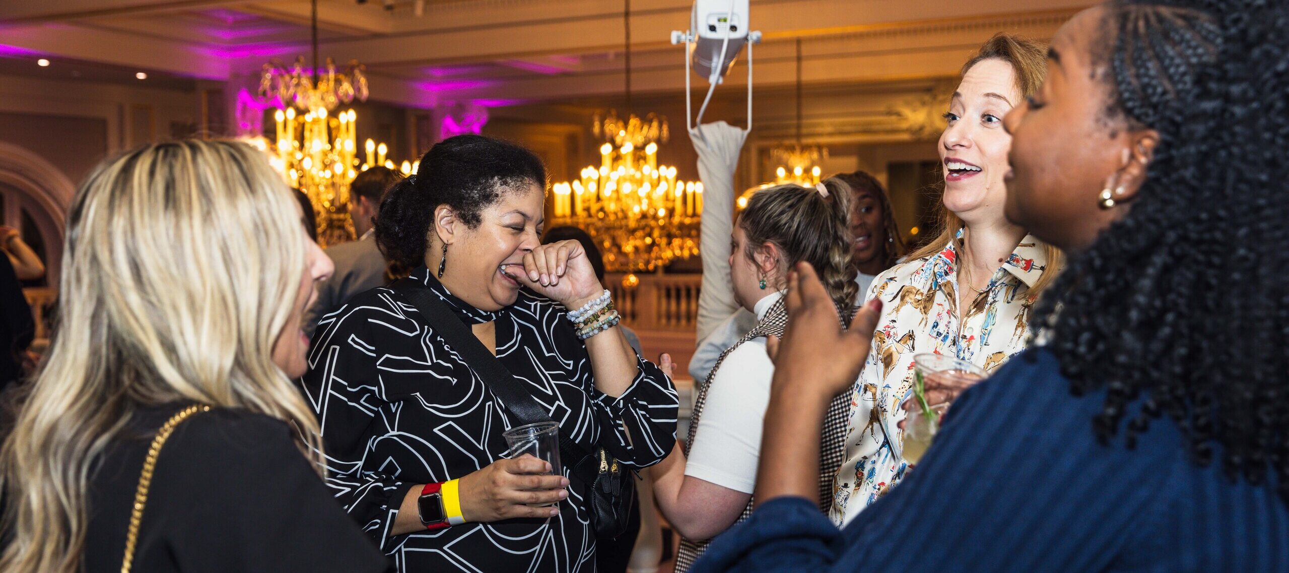 A group of people are socializing and laughing in an elegant room with chandeliers. They appear to be enjoying a lively conversation. One person holds a drink, and the atmosphere seems joyful and relaxed.