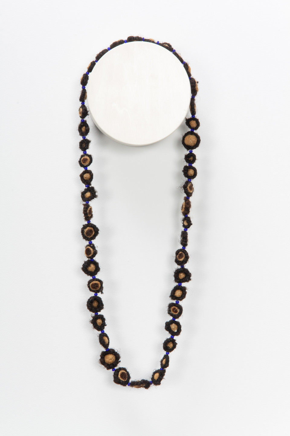 A necklace made from coiled, balled, and twisted dark human hair hangs on the wall. The hair is wrapped around small beads.
